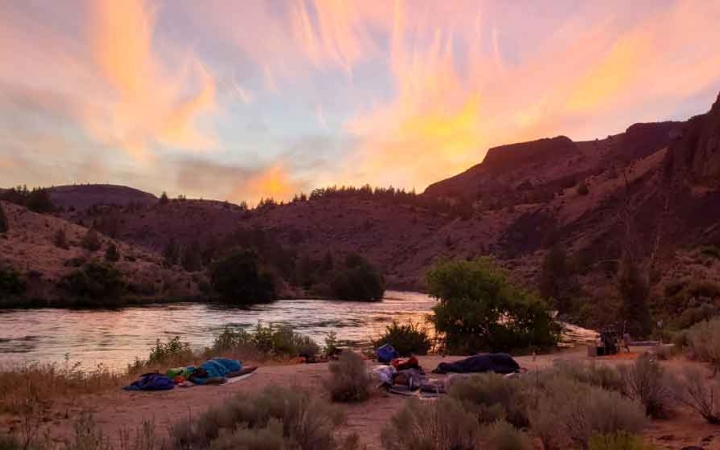 the sun rises behind a ridge in the background of a campsite along a river on an outward bound trip